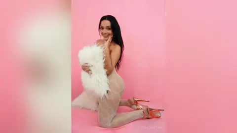 Smiling woman in a white faux fur coat and patterned tights posing against a pink background.
