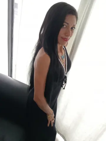 Elegant woman in a black sleeveless dress and necklace posing by a bright window in a serene indoor setting.