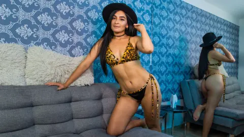 Confident woman in leopard print bikini and black hat posing on a gray sofa against blue damask wallpaper.