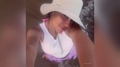Smiling woman in a white tank top and bucket hat enjoying a sunny day at the beach.