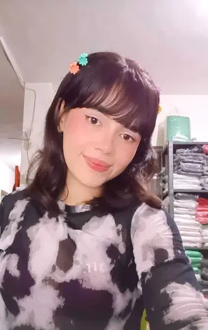 Smiling young woman with dark bangs wearing a black and white patterned mesh top in a cozy indoor setting.