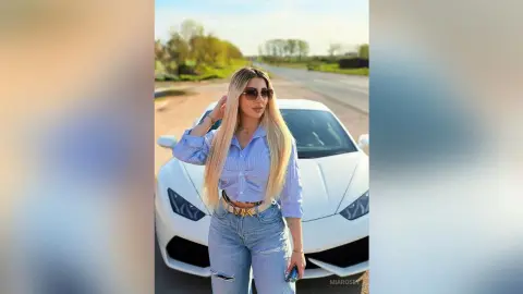 Stylish blonde woman in blue striped shirt and jeans posing confidently in front of a white luxury sports car.