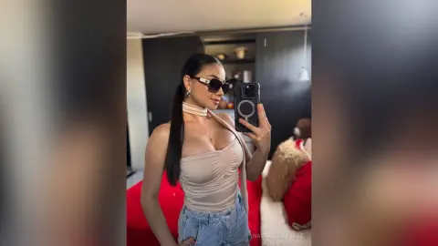Stylish woman in a beige bodysuit and sunglasses taking a mirror selfie in a modern bedroom.