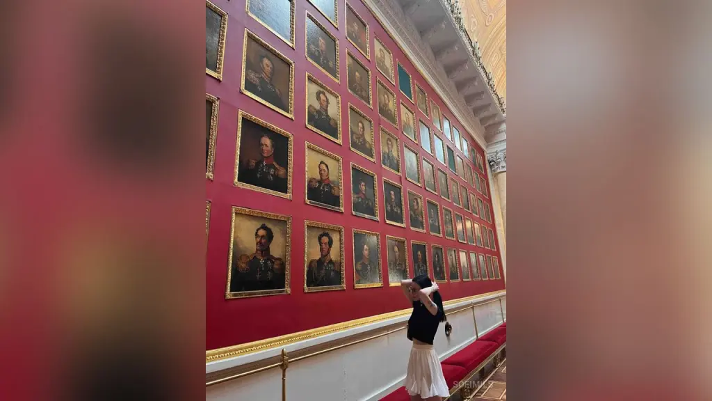 SofiMils β SofiMils stands in a grand gallery, looking at a large wall of framed portraits hanging above a red velvet bench.