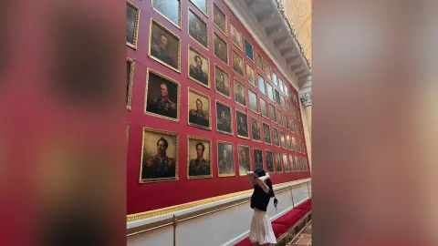 Young woman in a black top and white skirt standing before a gallery wall of vintage framed portraits.