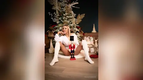 Blonde woman in white loungewear sitting by a Christmas tree with a nutcracker in a festive holiday setting.