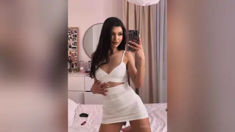 Young woman in a white ruched mini dress posing for a mirror selfie in a bright bedroom.