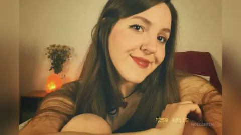 Smiling young woman with long brown hair wearing a black mesh top in a cozy, dimly lit room.