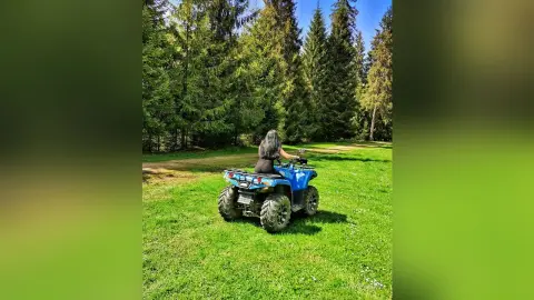 AnnaBanks with long dark hair sits on a blue ATV in a sunny, grassy clearing surrounded by a dense pine forest.
