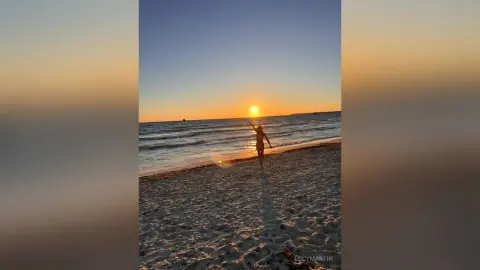 LucyMartin stands on a sandy beach at sunset, raising her arms toward the sun over the ocean waves.