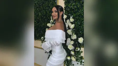 AnnaKarev poses in a tight white off-the-shoulder dress against a wall of white roses.