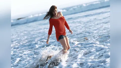 Young woman in a red knit top and denim shorts playing in ocean waves on a sunny beach.