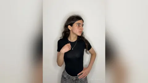 FondaYarbro — FondaYarbro wears a tight black t-shirt and grey jeans while standing against a white wall.