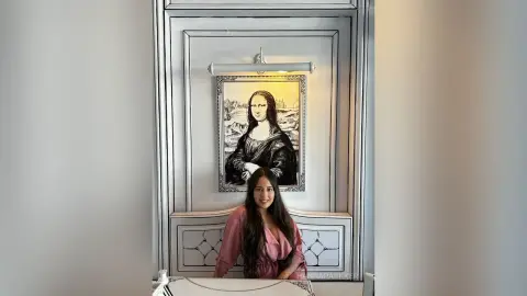Smiling woman in a pink blouse sitting in front of a Mona Lisa art piece in a modern white interior.