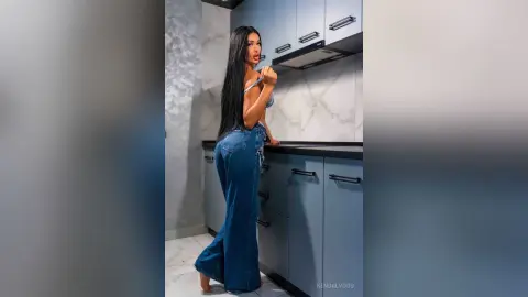 Woman in denim flared jeans and blue top posing in a modern marble kitchen.