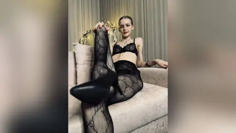 Elegant woman in black lace lingerie and patterned tights relaxing on a beige sofa in a moody indoor setting.