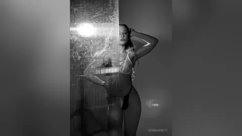 Moody black and white portrait of a woman in white lingerie posing behind a textured glass partition.
