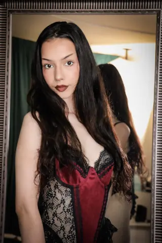 Young woman with long dark hair wearing a red and black lace corset in a moody indoor setting.