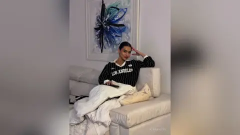 Stylish woman in a black Los Angeles sweatshirt and white pants relaxing on a white sofa in a modern room.