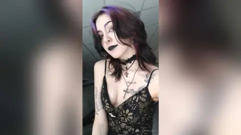 LorenLoiss poses in a black lace camisole with gold patterns, wearing black lipstick and a black choker with a cross pendant.