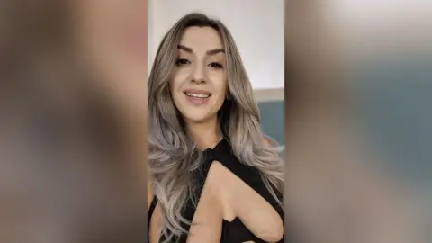 Smiling woman with long ash-blonde hair wearing a stylish black halter top in a bright indoor setting.