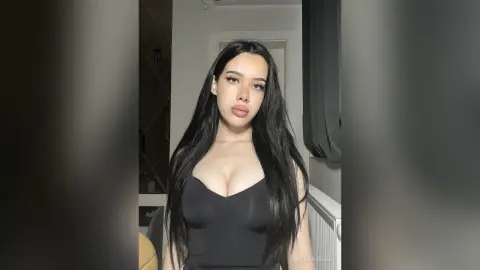 Young woman with long dark hair wearing a black tank top in a moody indoor setting.