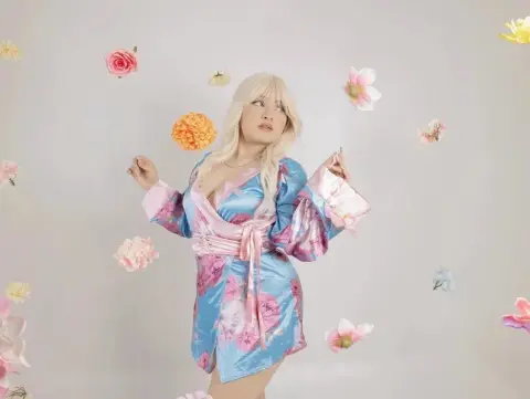 Blonde woman in a pink and blue floral satin robe posing amidst floating flowers in a dreamy studio setting.