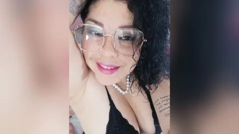 Smiling woman with curly hair and glasses wearing a black lace top in a bright indoor setting.