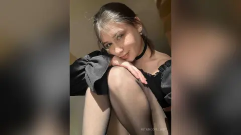 RitaMoonllight wearing a black top and choker, posing with her knees up and hands near her face in soft sunlight.