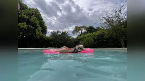 Woman in orange bikini relaxing on a pink floatie in a swimming pool under a cloudy sky.