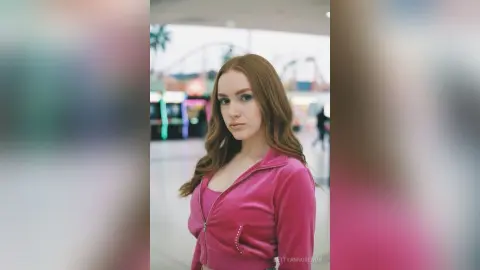 Young woman with long wavy red hair wearing a pink zip-up jacket at a bright amusement park.