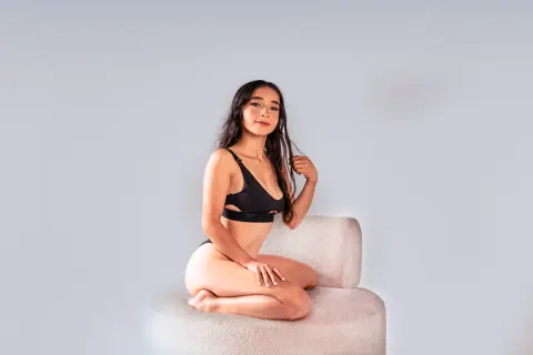 Young woman in black bikini sitting on a white chair against a plain background with a serene mood.