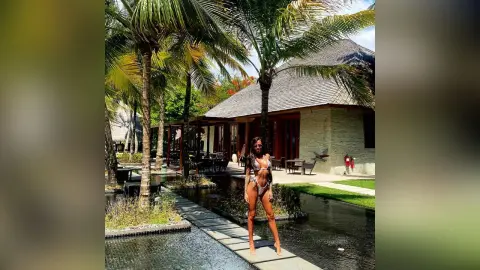 Woman in a white bikini standing by a tropical resort pond surrounded by palm trees on a sunny day.