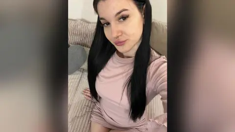 Young woman with long dark hair wearing a pink ribbed long-sleeve top sitting on a beige sofa in a cozy room.