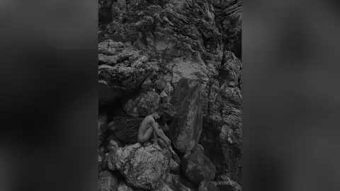 Nude woman sitting amidst rugged dark rocks in a moody, cinematic black and white nature setting.