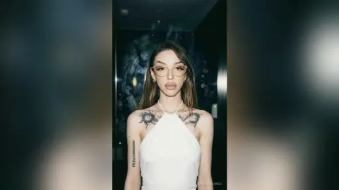 Young woman with tattoos in a white halter top posing confidently in a dark, moody indoor setting.