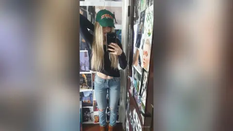 Young woman in a black long-sleeve shirt, blue jeans, and green baseball cap taking a casual mirror selfie.