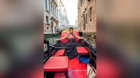 MissyJolie sits in a gondola wearing a black lace dress with a low-cut neckline, posing against a red backrest on the water.