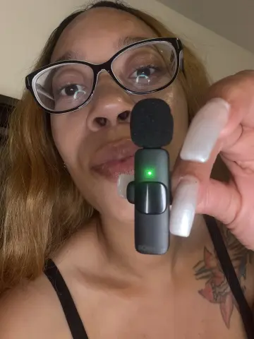 LexBright holds a small black microphone near her face, wearing black glasses and a black tank top, showing her light brown hair and chest tattoo.
