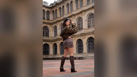 Stylish woman in a brown faux fur jacket and mini skirt posing in a historic courtyard setting.