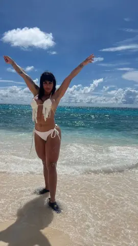 Cheerful woman in a white crochet bikini standing on a sunny tropical beach with turquoise ocean waves.