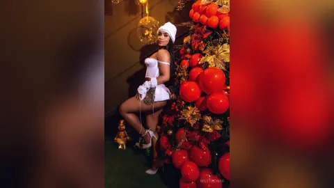 Glamorous woman in a white satin outfit posing by a festive red Christmas tree in a warm holiday setting.