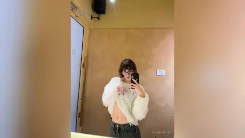 Young woman in a white fuzzy sweater and denim jeans taking a mirror selfie in a modern bathroom.