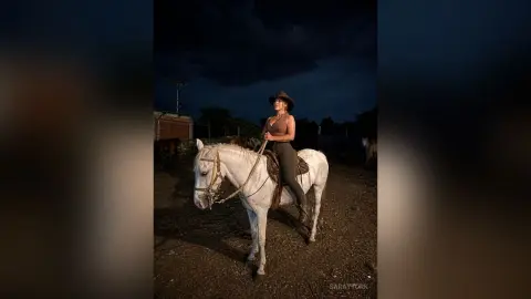 Woman in equestrian attire riding a white horse in a dark ranch setting under a moody twilight sky.