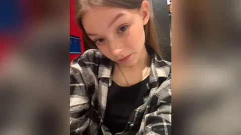 Young woman in a black top and grey plaid flannel shirt posing in a casual indoor setting.