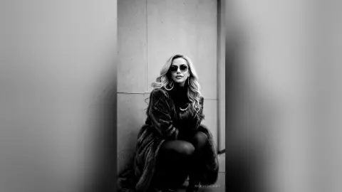 Stylish blonde woman in sunglasses and a dark faux fur coat posing against a concrete wall in a moody black and white portrait.