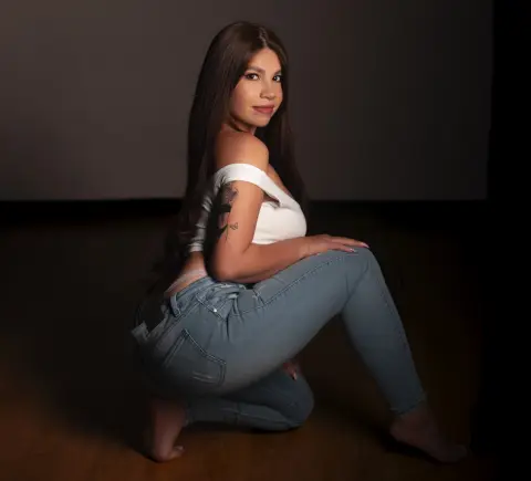 Young woman in white off-the-shoulder top and blue jeans posing in a studio setting with a moody atmosphere.