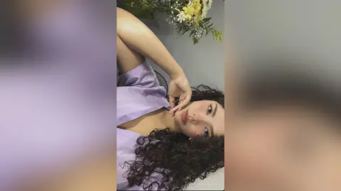 Smiling young woman with curly hair wearing a lavender blouse in a bright, floral indoor setting.