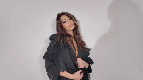BlairBlanche poses in a black ruffled blazer with her long brown hair flowing against a plain grey background.
