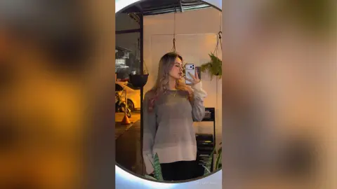 HillaryLewis poses for a mirror selfie wearing a long-sleeved, light grey ombre sweater and black pants, with her long blonde hair down.